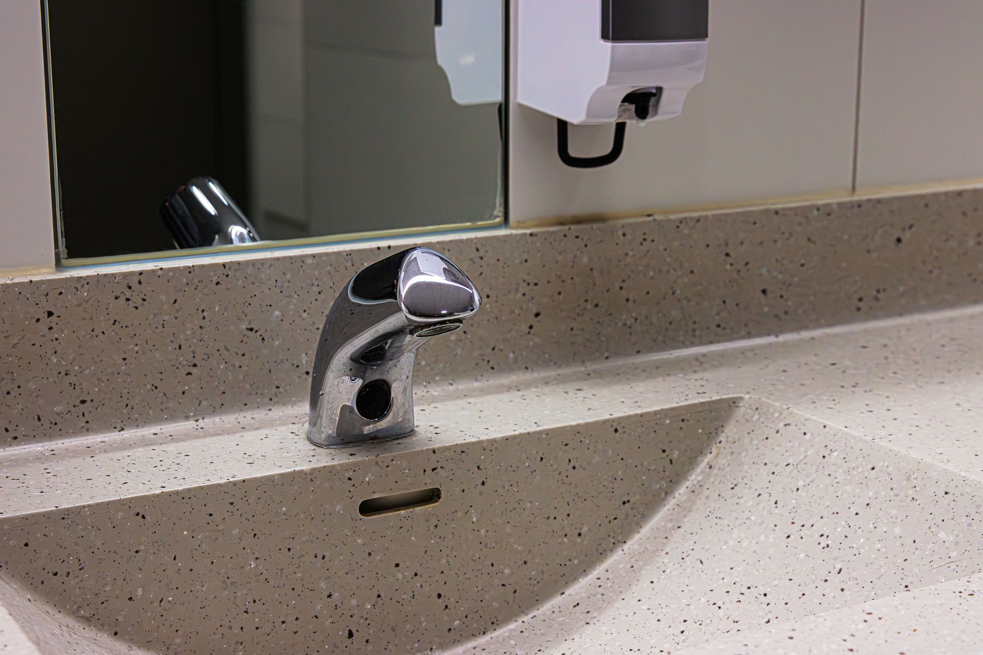 Modern sensor faucet mounted on a gray stone basin, mirror, and soap dispenser in public restroom.